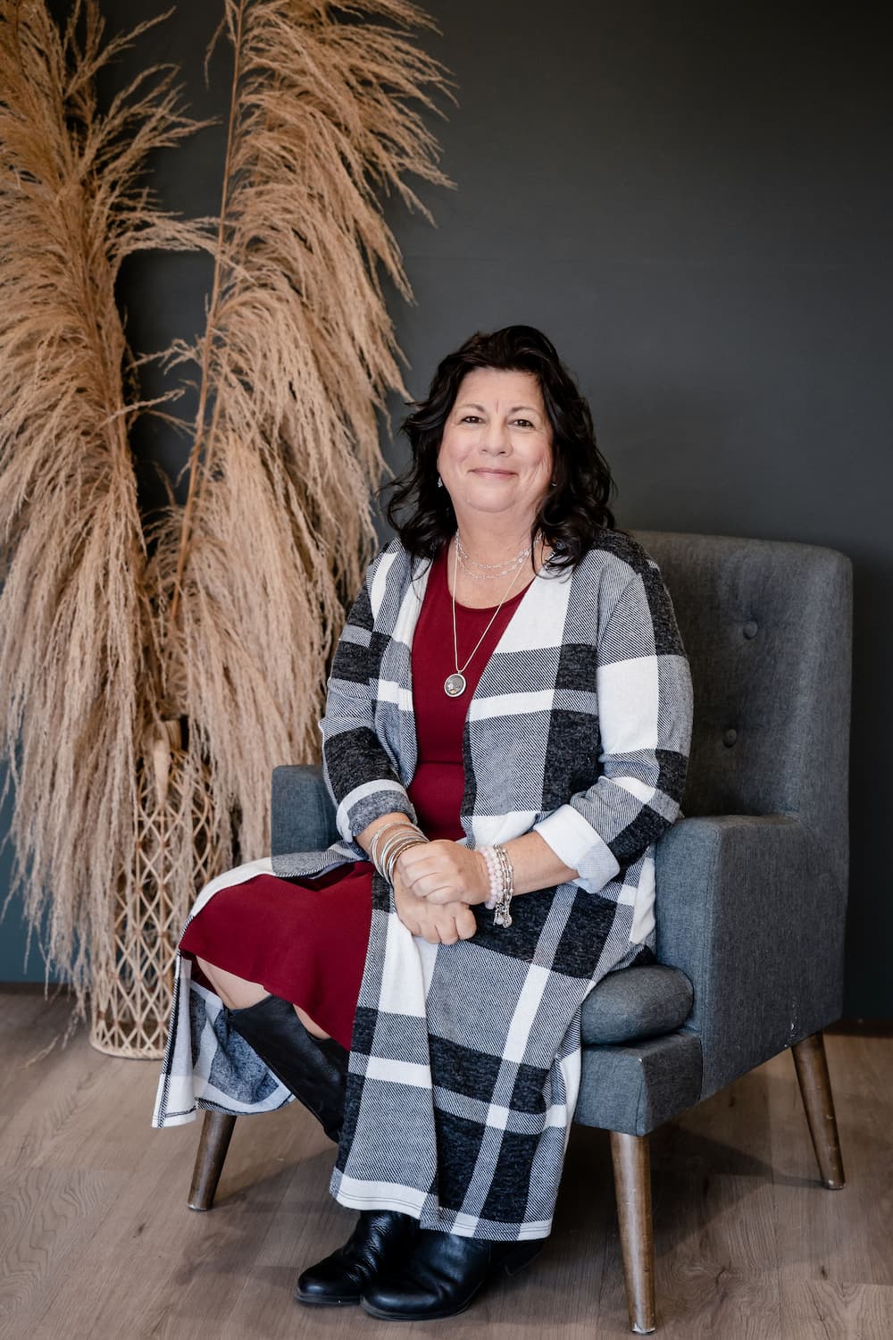 A woman with dark hair wearing a red dress and a black-and-white plaid cardigan sits on a gray armchair, smiling. Tall dried pampas grass decorates the background against a dark wall.