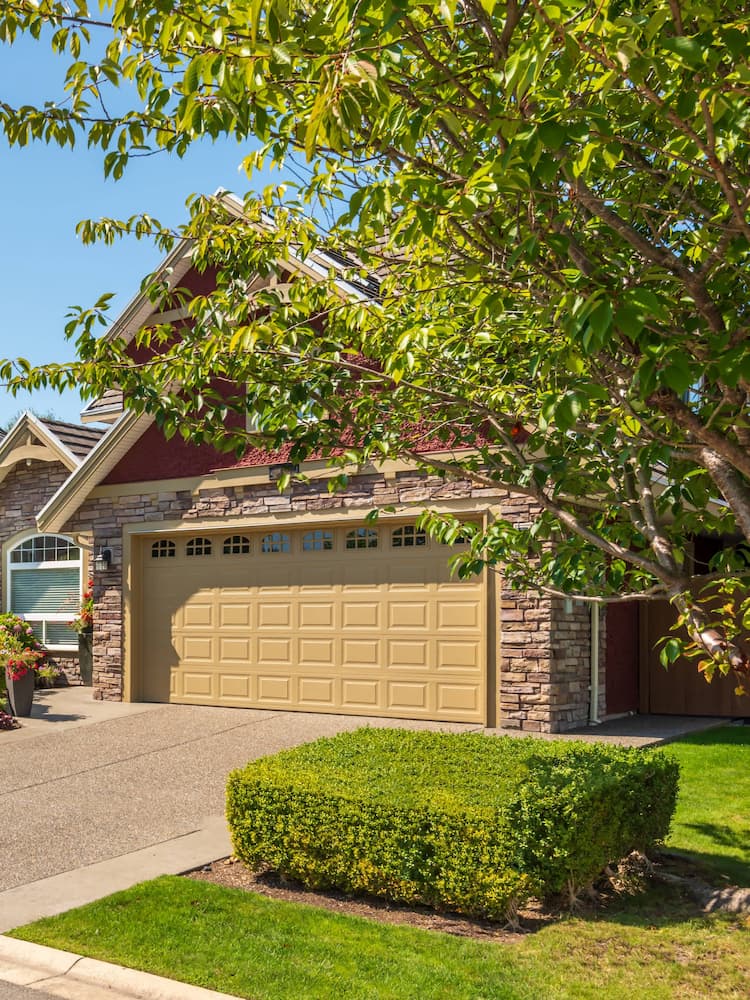 A suburban house with stone and red siding, featuring a tan double garage door, well-kept shrubs, and a leafy tree in the front yard on a sunny day.