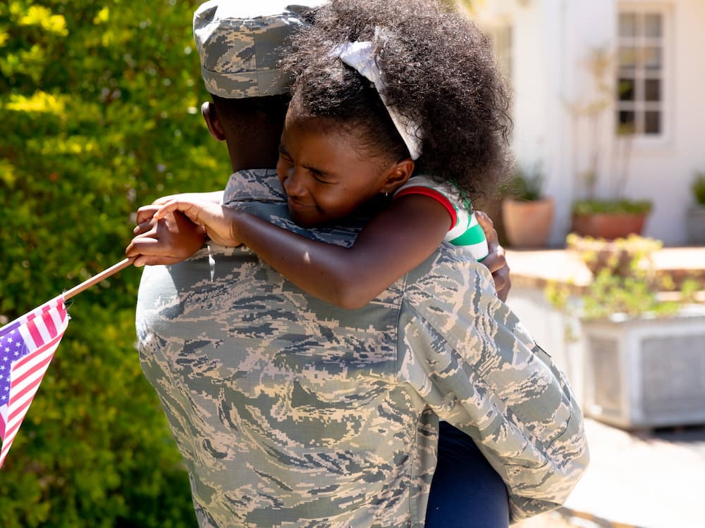 A soldier in uniform hugs a young girl tightly outside a house. The girl, smiling with eyes closed, holds a small American flag as she clings to the soldier’s neck, expressing joy and affection.