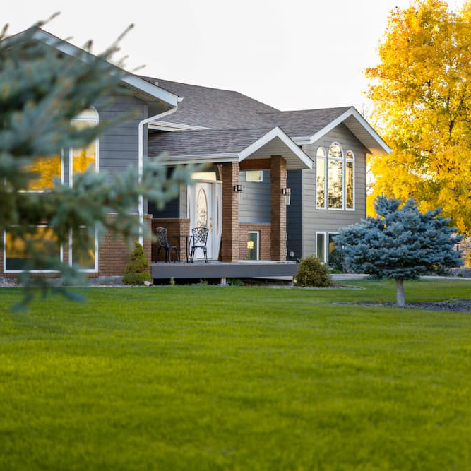 A modern house with large windows and a front porch sits behind a well-kept lawn, surrounded by trees, including one with bright yellow autumn leaves.