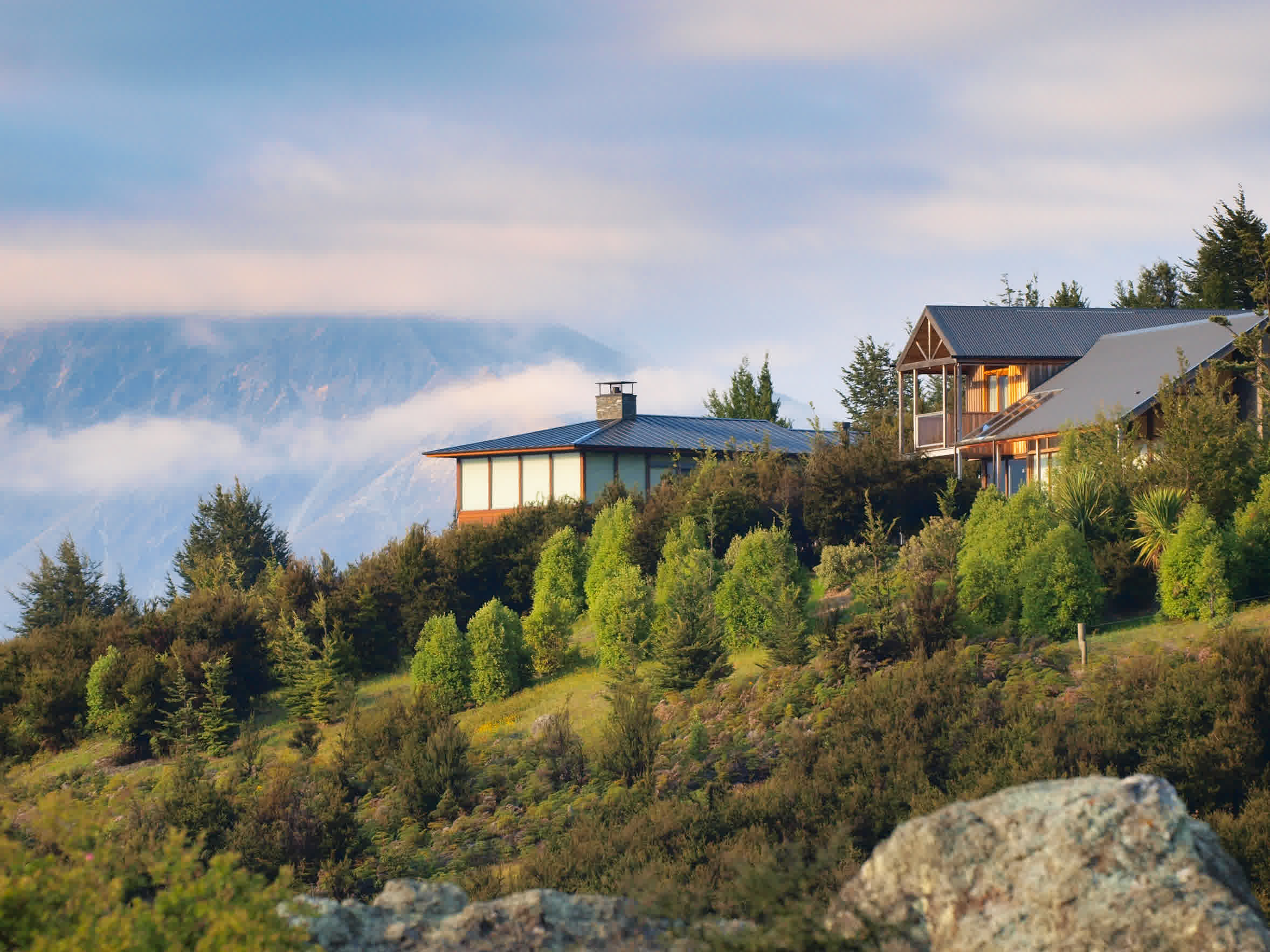 A hillside home surrounded by lush green trees and bushes, with mountains in the background partially covered by clouds, under soft, early morning or evening light.