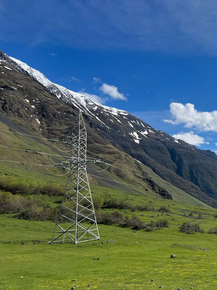 A metal electricity pylon stands on a green grassy field, with a backdrop of snow-capped mountains and a blue sky with scattered clouds.
