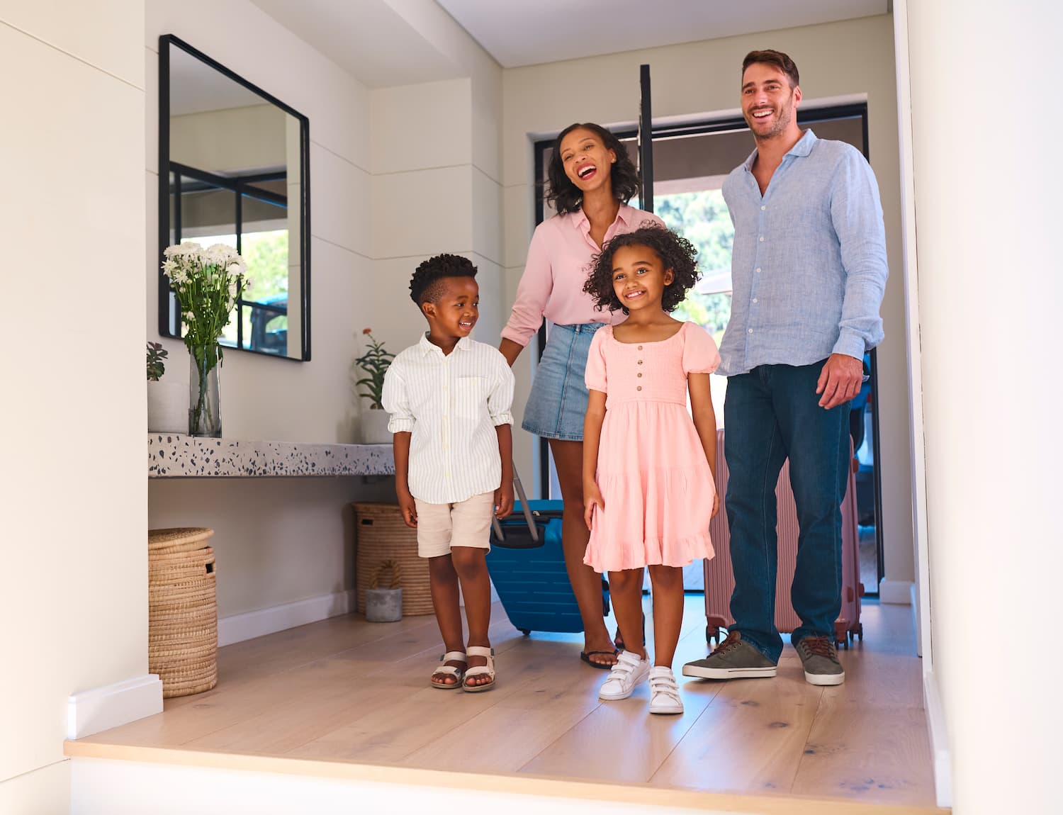A smiling family of four, two adults and two children, stands in the entryway of a bright, modern home, holding luggage, appearing happy and ready for a trip or arrival.