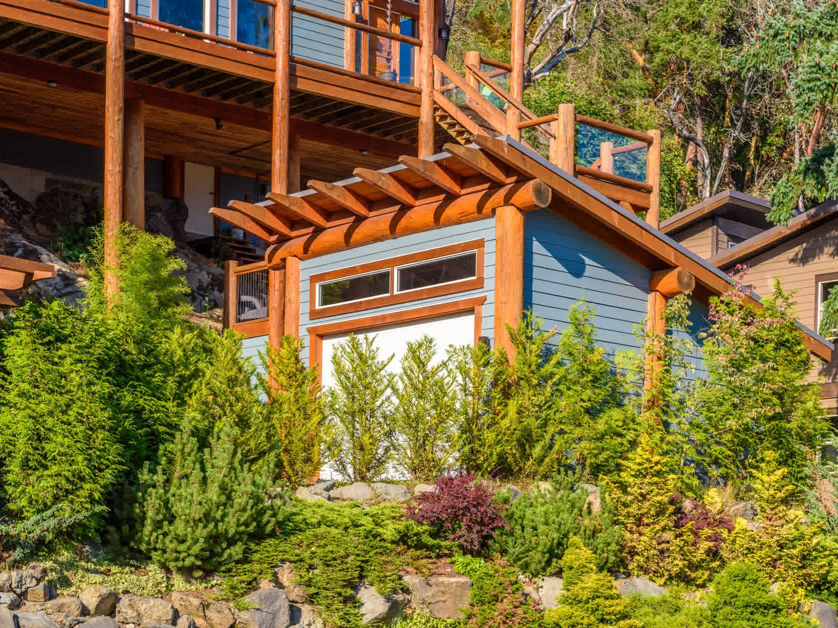 A small modern shed with blue siding and wooden trim sits among green shrubs and trees, with a large deck and house visible above and behind it.