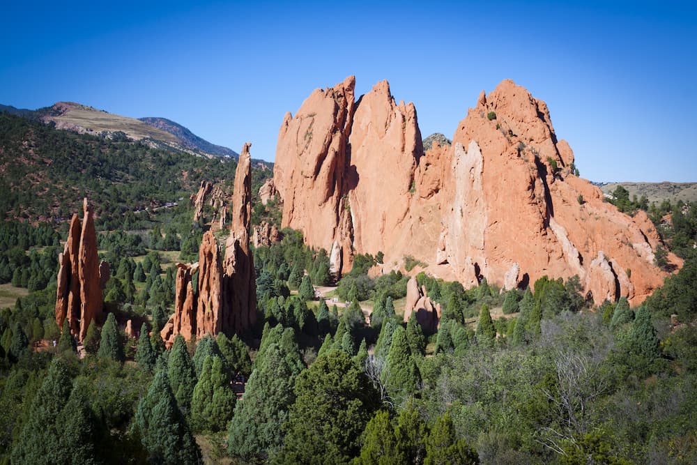 Towering red sandstone rock formations rise above green trees under a clear blue sky at Garden of the Gods in Colorado Springs, with distant hills visible in the background.