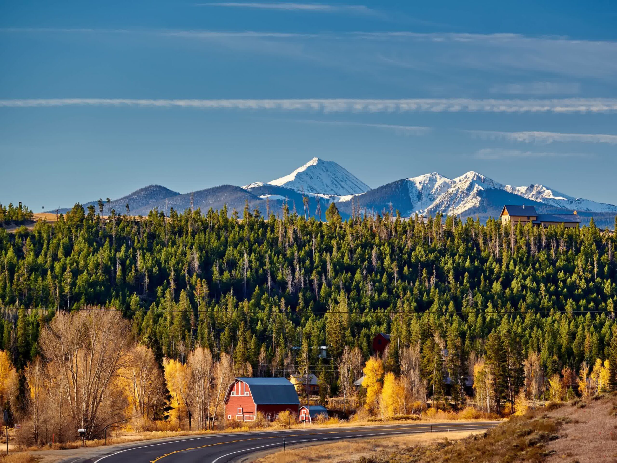 A winding road passes by a red barn and autumn trees, with a dense evergreen forest and snow-capped mountains in the background under a clear blue sky.