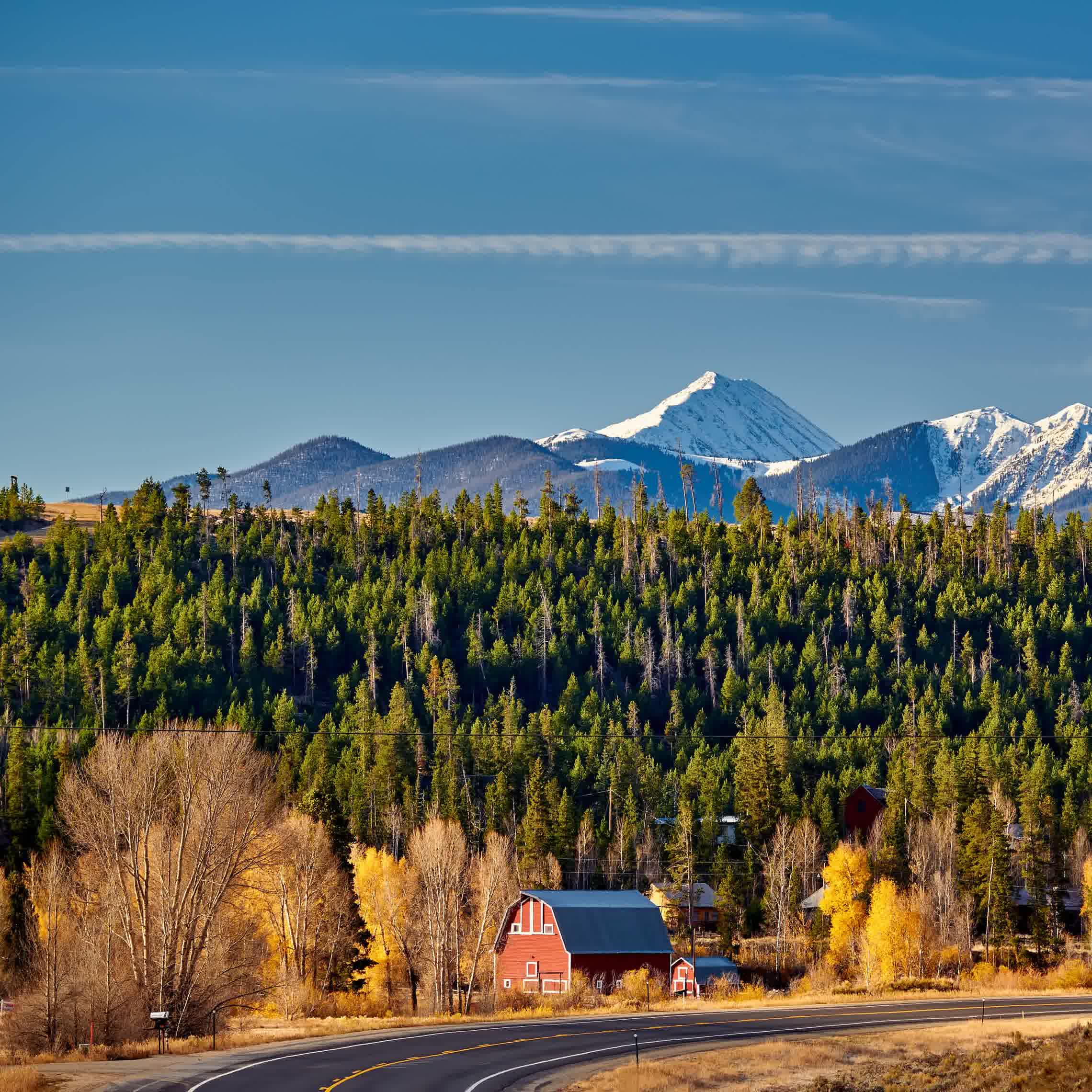Curving road passes a red barn surrounded by trees with autumn foliage, dense pine forest, and snow-capped mountains under a clear blue sky in the background.