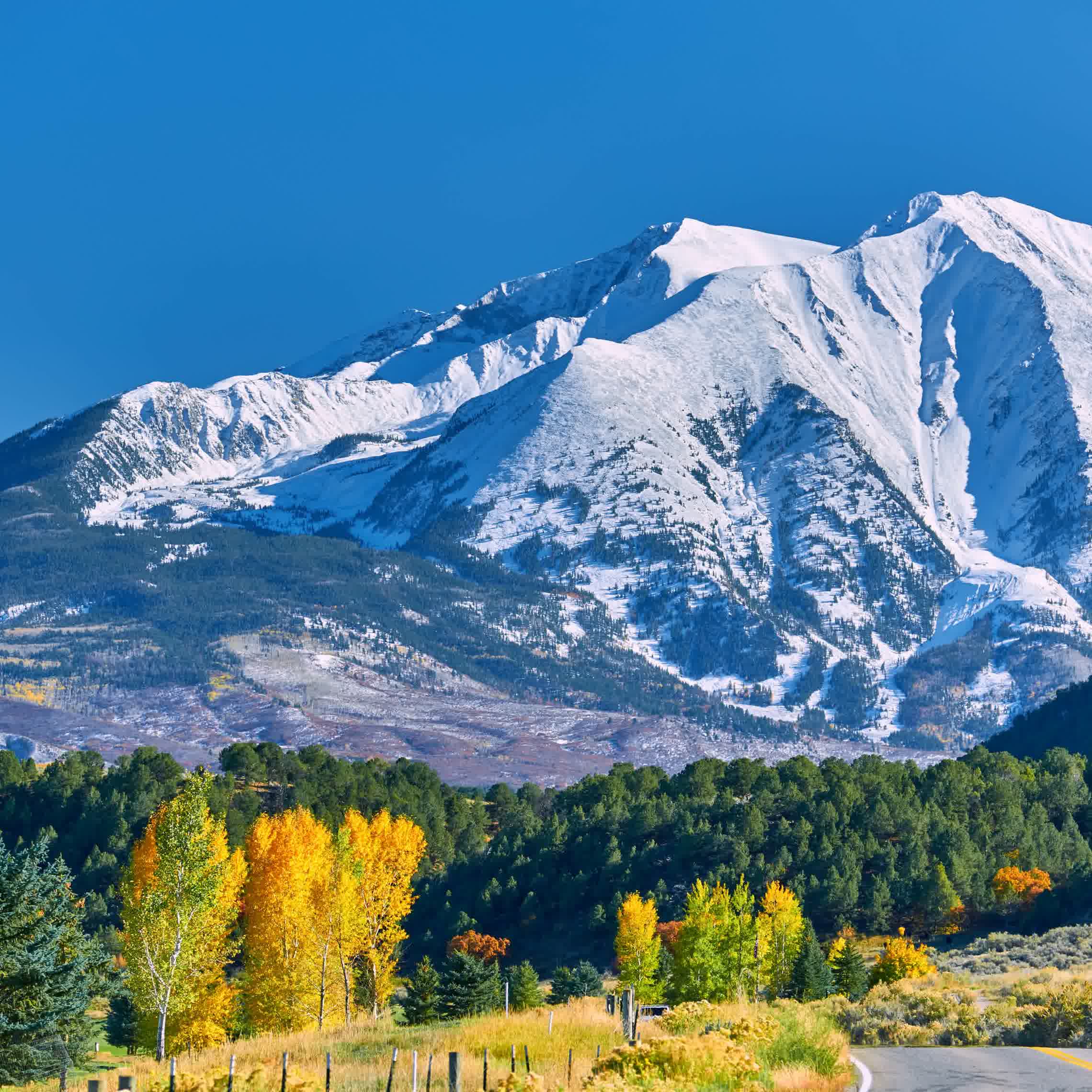 A scenic view of a snow-capped mountain under a clear blue sky, with colorful autumn trees and a winding road in the foreground.