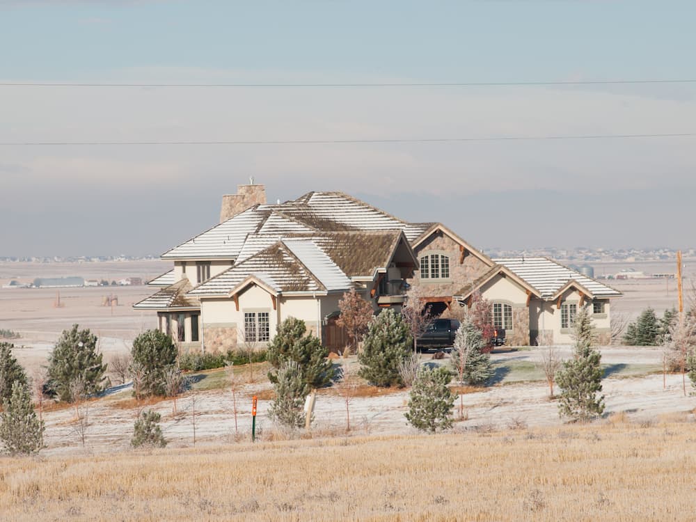 A large, modern house with a snow-dusted roof sits on a spacious property surrounded by evergreen trees and a frosty landscape under a clear sky.