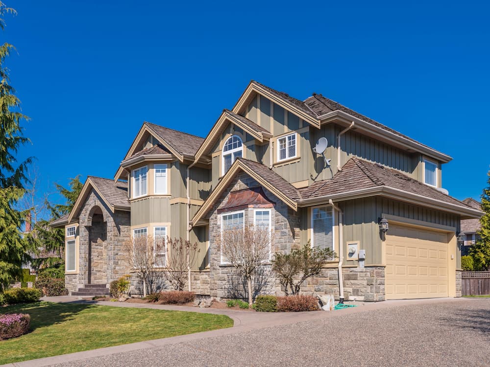 Two-story suburban house with beige siding, stone accents, multiple gabled roofs, and a double garage. Well-maintained landscaping and clear blue sky in the background.