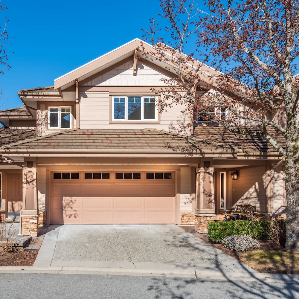 Two-story suburban house with tan siding, brown shingled roof, double garage door, upper and lower windows, and a tree with bare branches in the front yard under a clear blue sky.
