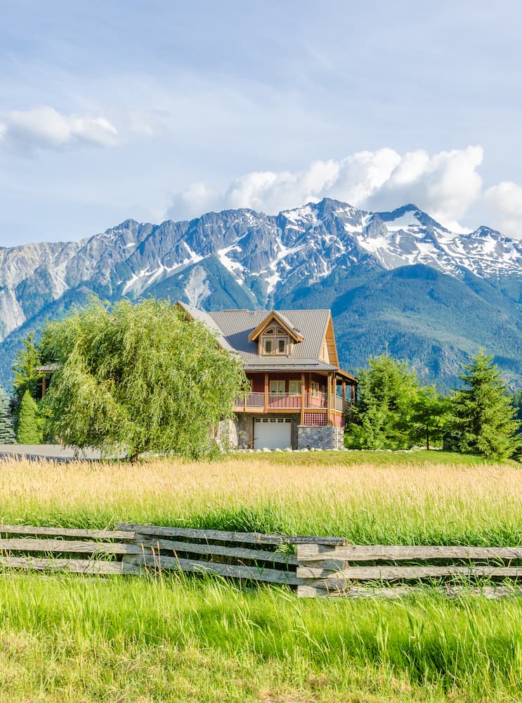 A wooden house with a balcony sits in a grassy field, surrounded by trees, with tall, snow-capped mountains in the background under a blue sky. A wooden fence runs along the foreground.