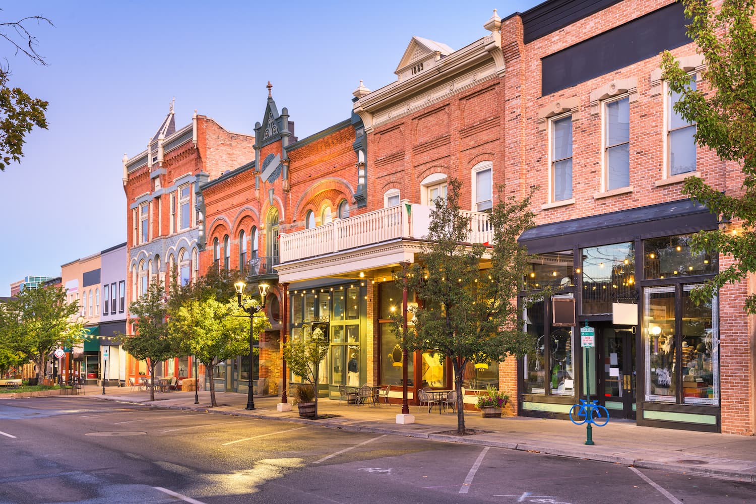 A quiet small-town street at dusk with historic brick buildings, large windows, trees lining the sidewalk, and soft streetlights illuminating the empty road.
