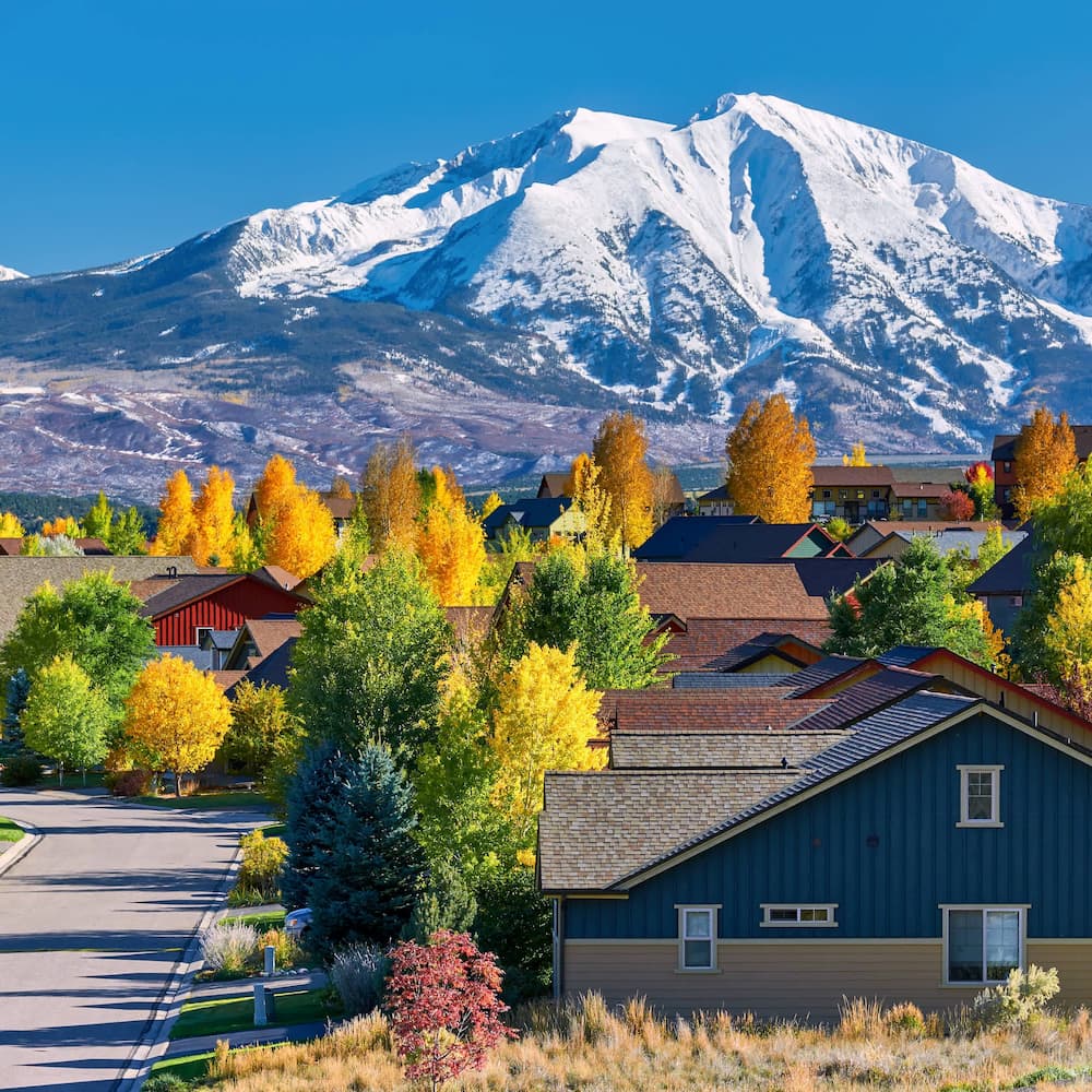 A suburban neighborhood with colorful autumn trees and houses, set against a backdrop of snow-capped mountains under a clear blue sky.