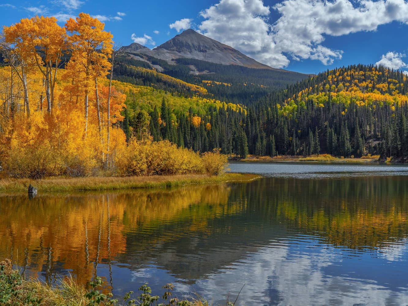 A serene lake reflects autumn trees with yellow and orange foliage, evergreen forest, and a mountain peak under a partly cloudy blue sky.
