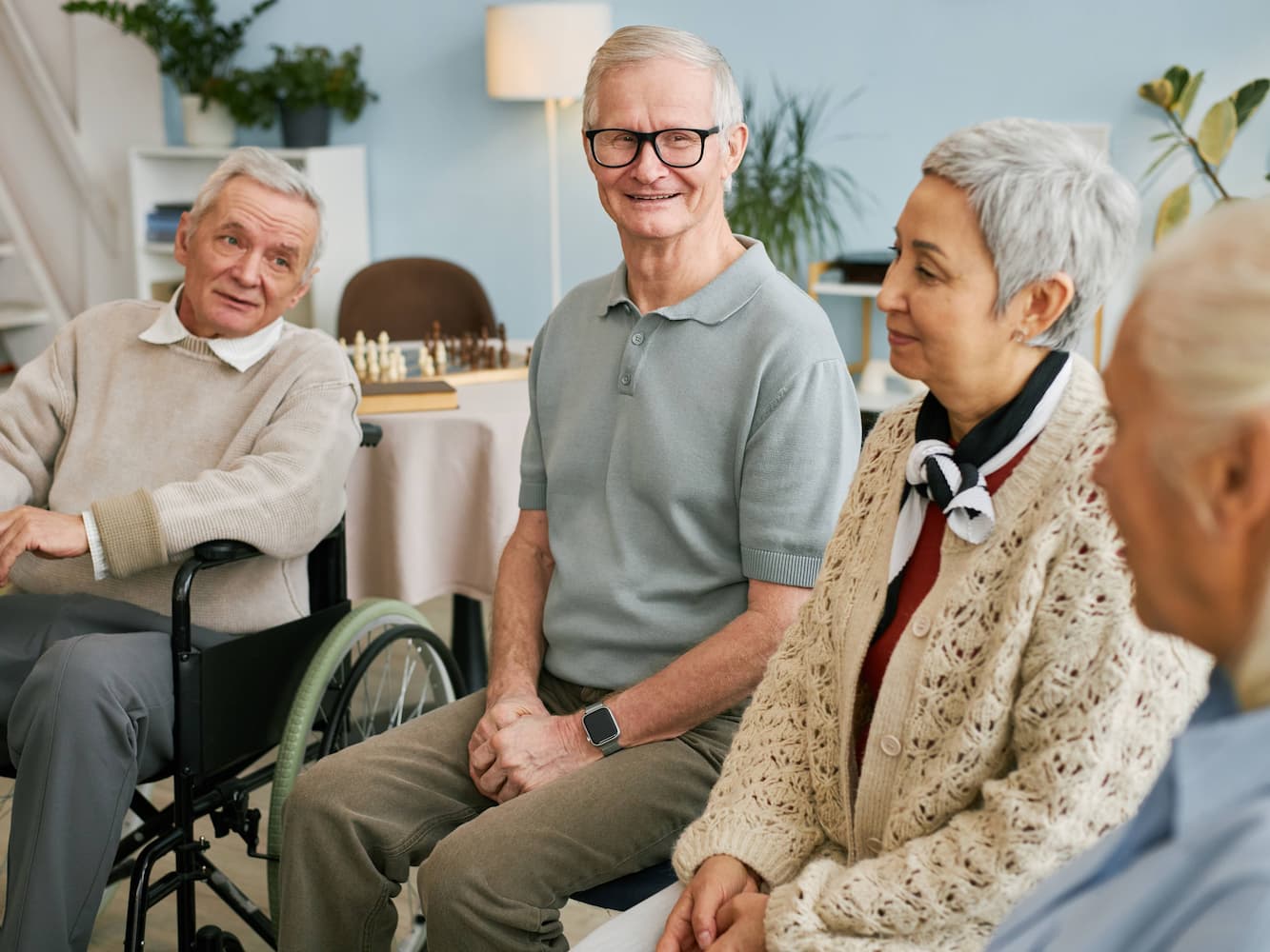 Four older adults sit together indoors, smiling and conversing. One person is in a wheelchair, while others sit on chairs. The room has soft lighting, plants, and a chessboard in the background, creating a warm, friendly atmosphere.