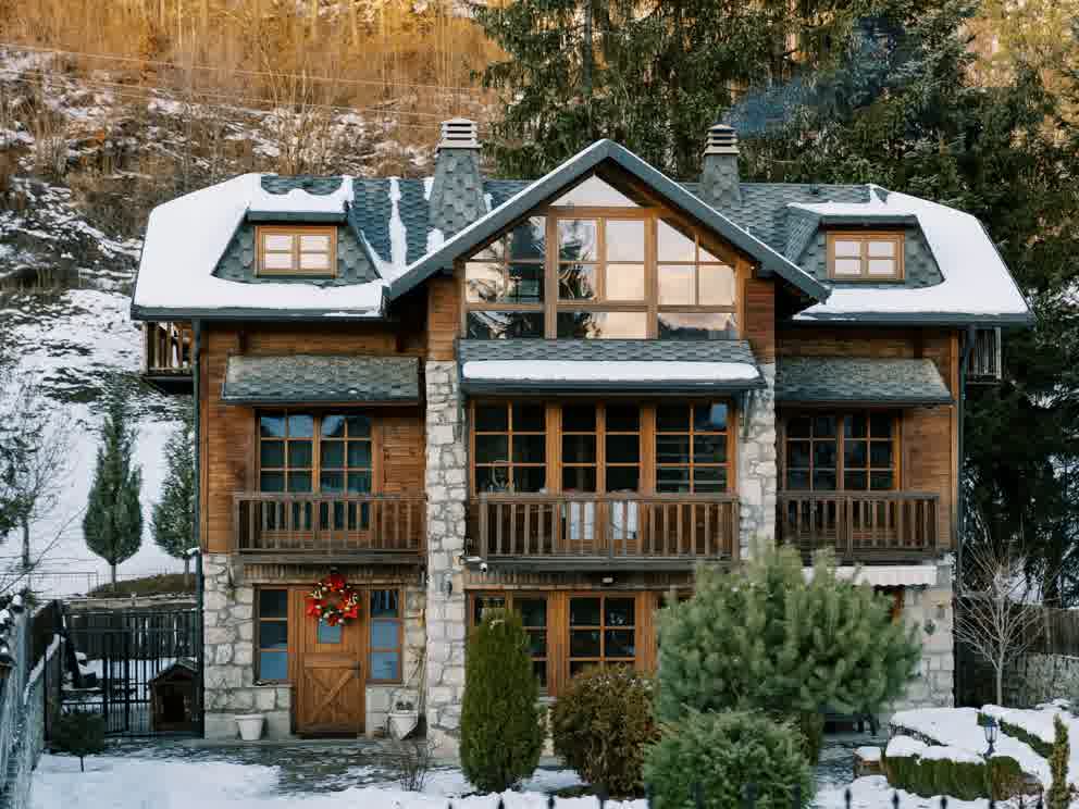 A cozy two-story wooden and stone house with snow on the roof, balconies, large windows, and a wreath on the front door, surrounded by trees and a snowy landscape.