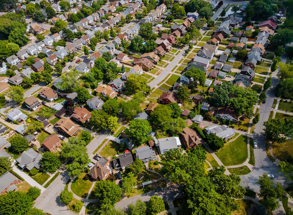 Aerial view of a suburban neighborhood with rows of houses, green lawns, and tree-lined streets laid out in a grid pattern on a sunny day.