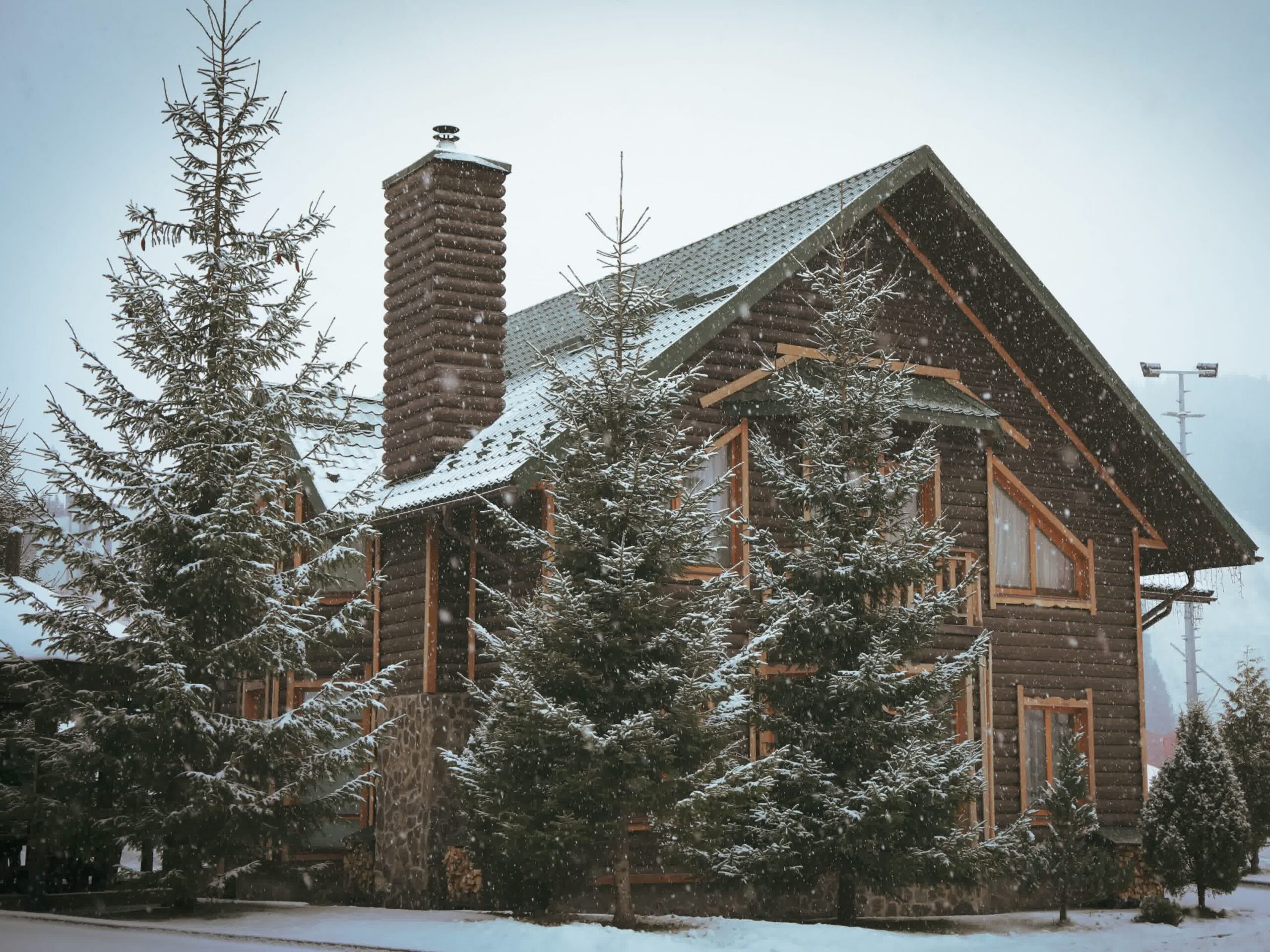 A cozy wooden cabin with a stone chimney is surrounded by snow-covered pine trees on a snowy winter day. Snowflakes are falling, creating a peaceful, festive atmosphere.