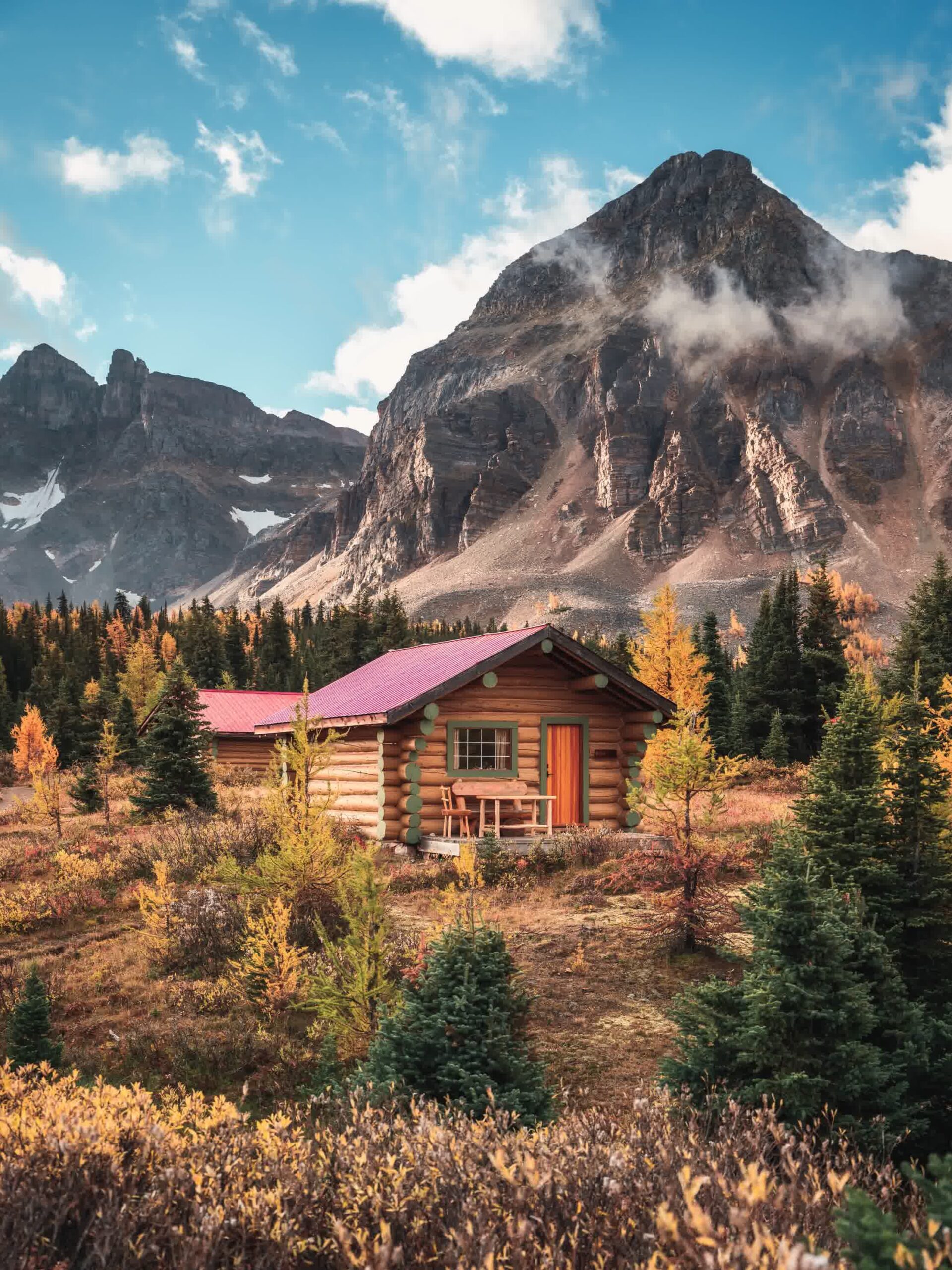A small log cabin with a red roof sits among evergreen trees and autumn foliage, with towering rocky mountains in the background under a partly cloudy sky.